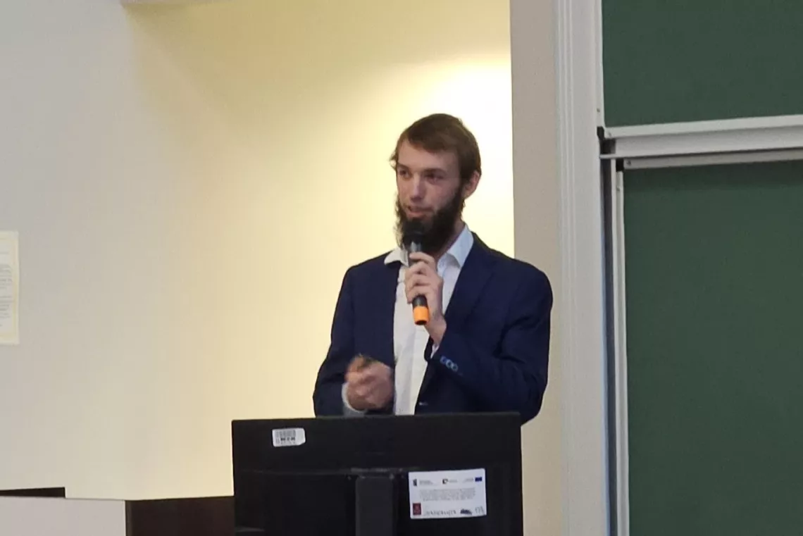 Młody mężczyzna z brodą przemawia, trzymając mikrofon w ręku