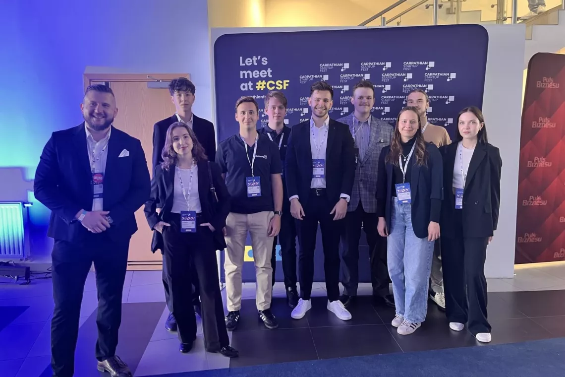 Grupa osób z identyfikatorami zawieszonymi na szyi stoi na tle banera organizatorów wydarzenia