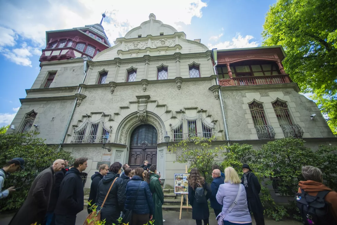 Grupa osób oglada zabytkową willę stojącą wśród zieleni