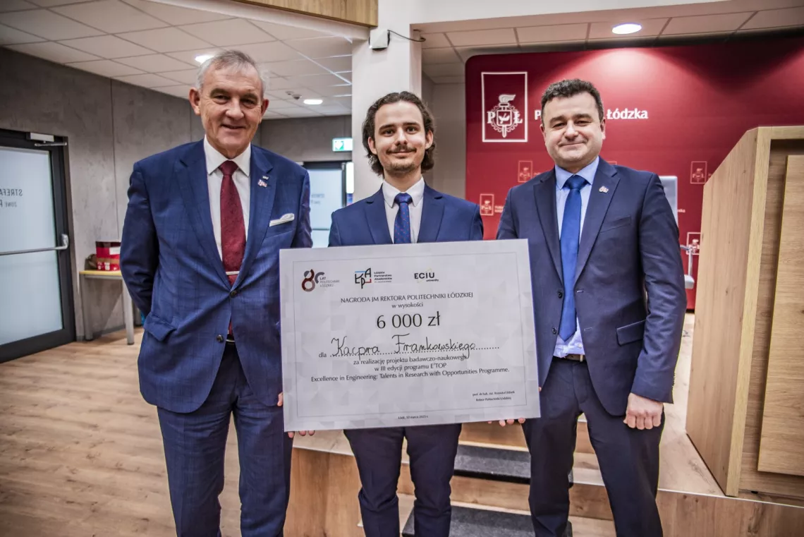 Młody człowiek stoi, trzymając czek w ręku, w środku, obok dwóch mężczyzn