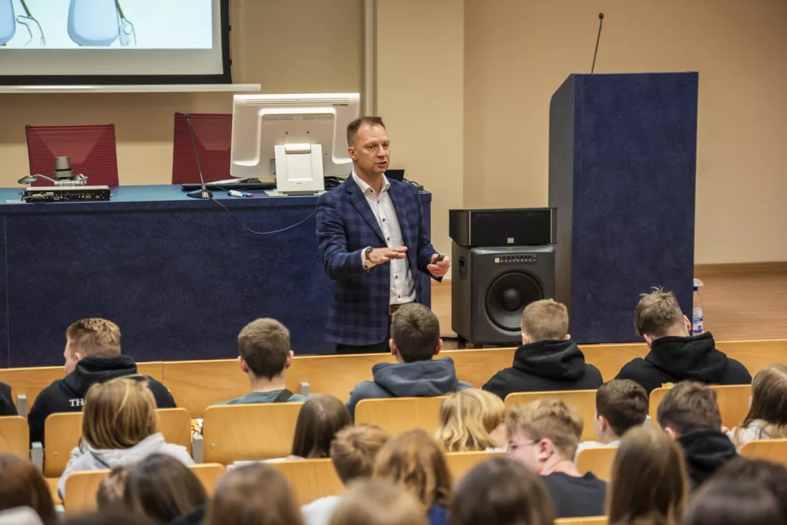 Osoba stojąca przed  salą wykładową, prowadząca prezentację. Prezenter znajduje się w pobliżu katedry. Młodzież siedzi w rzędach i uważnie słucha wykładu.