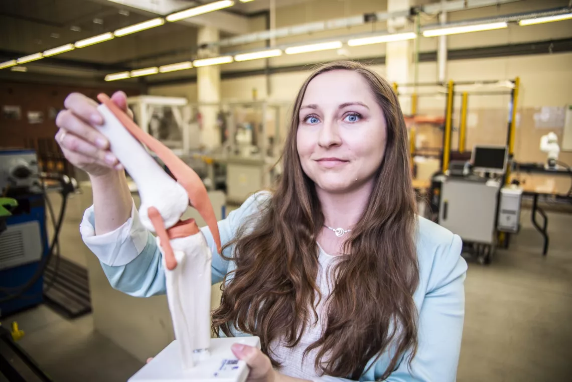 Osoba w laboratorium ltrzymająca model ludzkiego stawu kolanowego. W tle znajdują się różne urządzenia i maszyny.