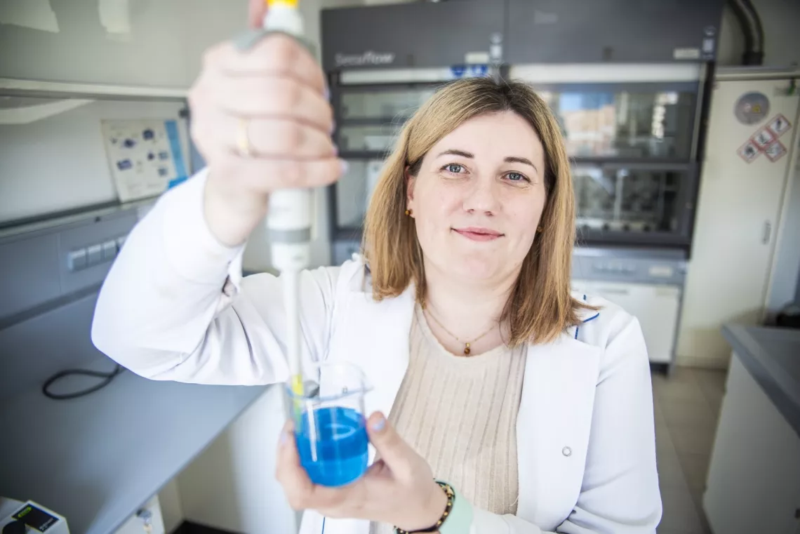 Osoba w laboratorium, która trzyma pipetę i dozuje niebieski płyn do zlewki.