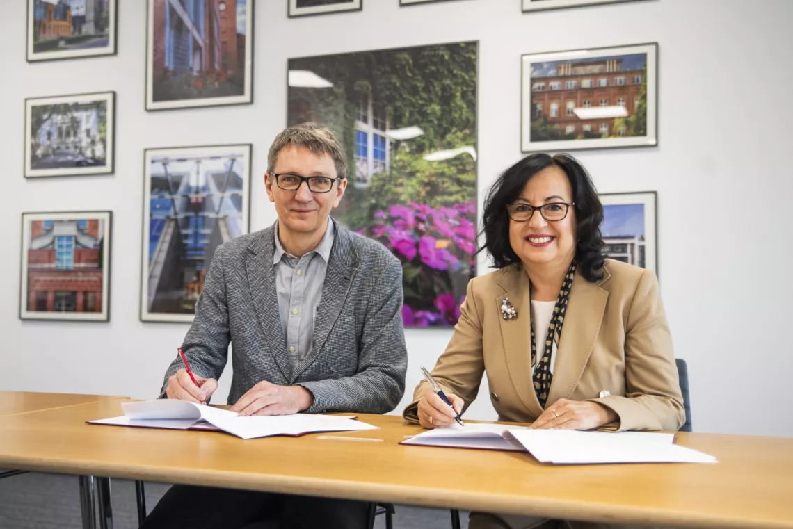 Dwie osoby przy stole podpisują dokumenty, siedząc przy stole. W tle biała ściana, na której wiszą zdjęcia oprawione w ramki.