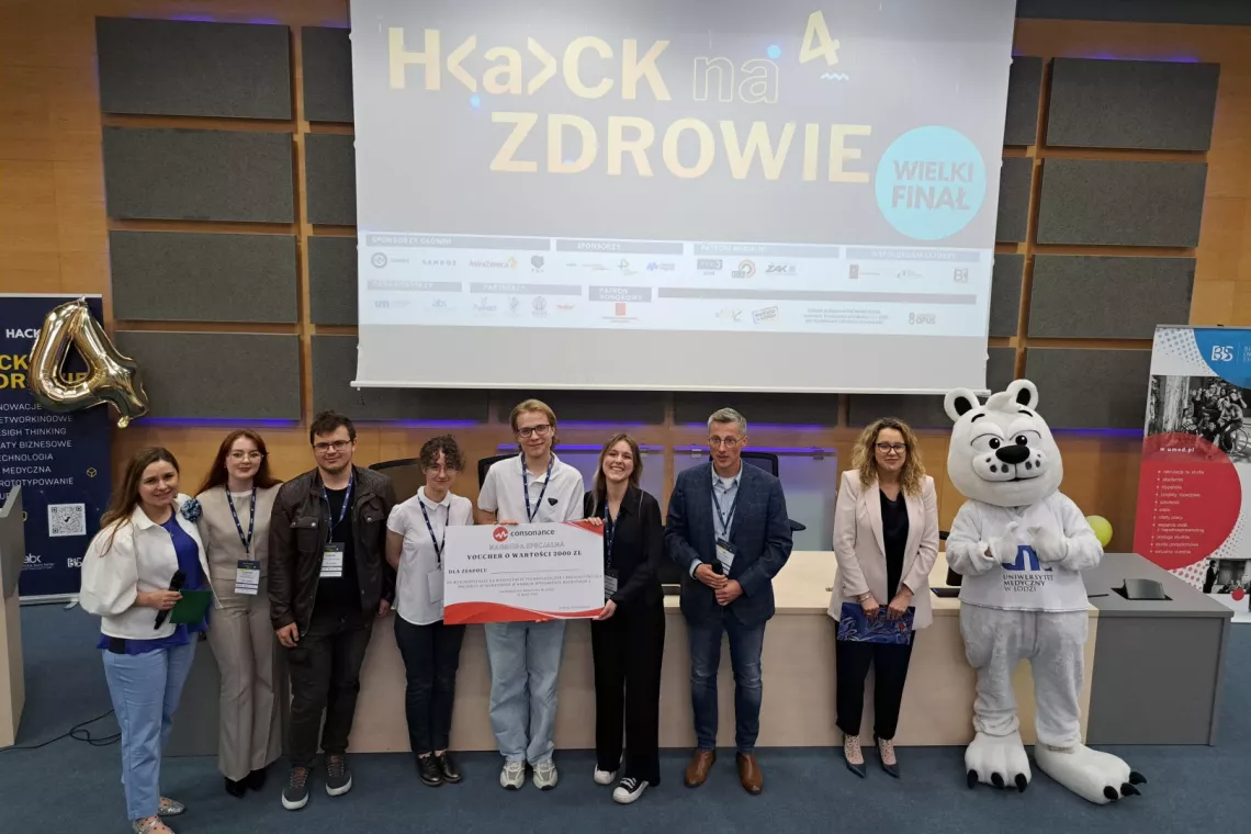 Osoby stoją przed ekranem z napisem Hack na zdrowie. Jedna z nich trzyma czek