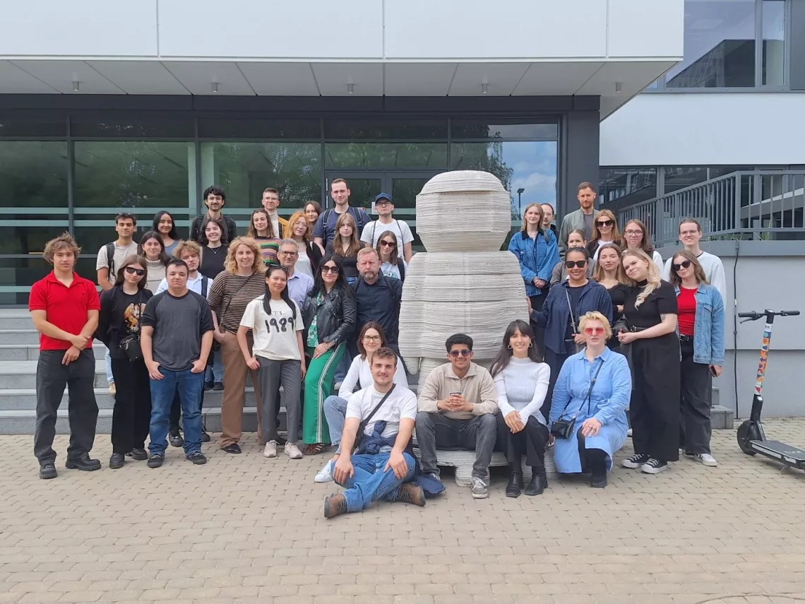 Liczna grupa studentów i opiekunów pozuje wspólnie przed szklanym wejściem do budynku i dużej rzeźby złożonej z poziomych warstw. Po prawej stronie widoczna hulajnoga elektryczna.