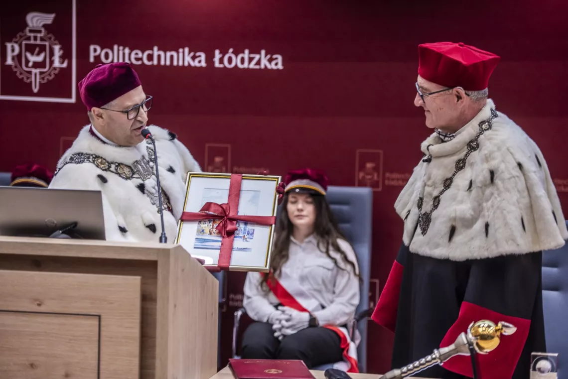 Osoba w rektorskim stroju przekazuje prezent-obraz drugiej osobie w rektorskim stroju. Z tyłu pod napisem Politechnika Łódzka siedzi osoba w czapce studenckiej, w bluzce przewiązanej szarfą.