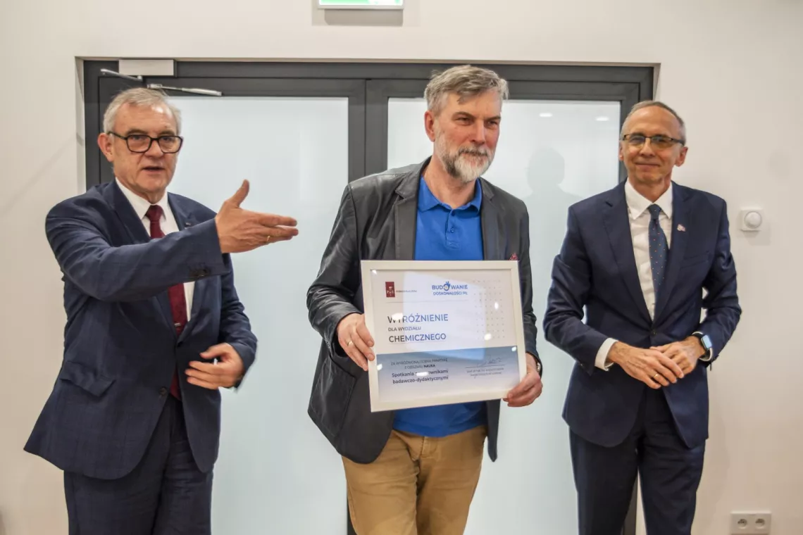 Trzech mężczyzn stoi, ten z lewej wskauje dłonią na stojącego w środku, który trzyma dyplom w ręku. Wszyscy trzej mają zadowolony wyraz twarzy.