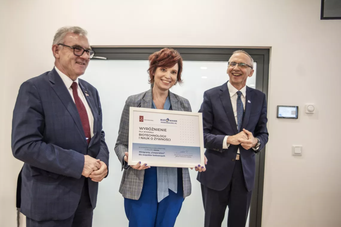 Trzy uśmiechnięte osoby, kobieta i dwóch mężczyzn stoją, kobieta prezentuje trzymany w ręku dyplom