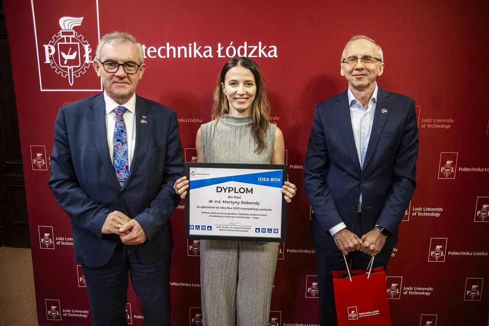 Trzy osoby stoją przed banerem, ta w śrdoku trzyma dyplom w ręku