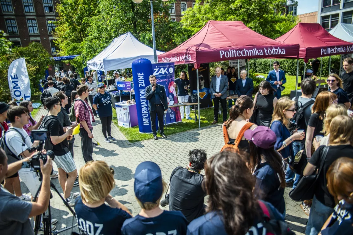 Mężczyzna z mikrofonem przemawia do grupy osób. Wszyscy stoją na świezym powietrzu, widać w tle zadaszone stoiska i banery reklamowe