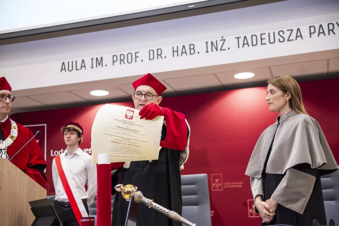 Mężczyzna w todze odczytuje tekst dyplomu z widocznym godłem Polski, stojącej obok  kobiecie w todze