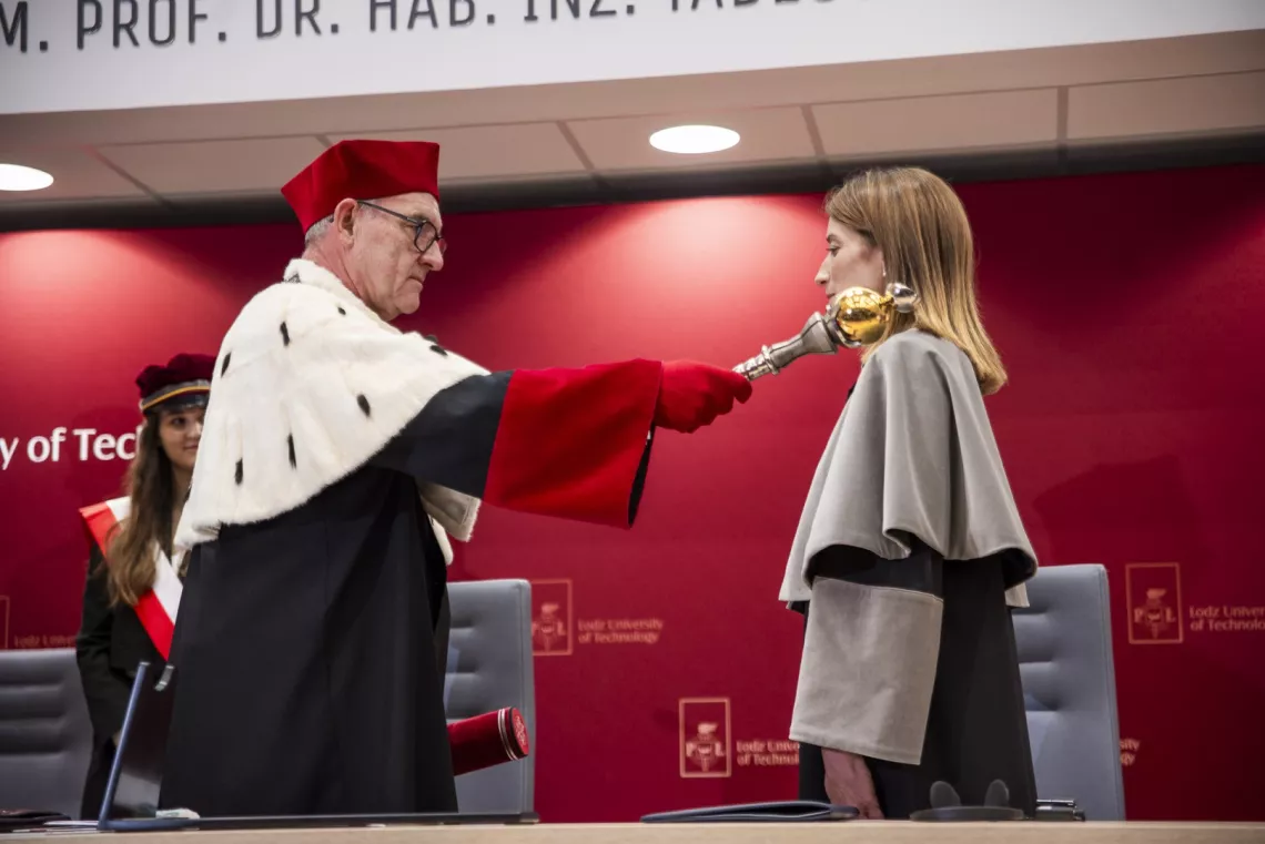 Mężczyzna w todze dotyka berłem ramienia stojącej przed nim kobiet w todze 