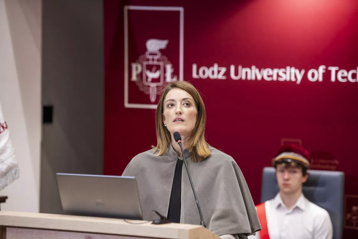 Kobieta w szarej todze wygłasza przemówienie przy mównicy, na której stoi laptop