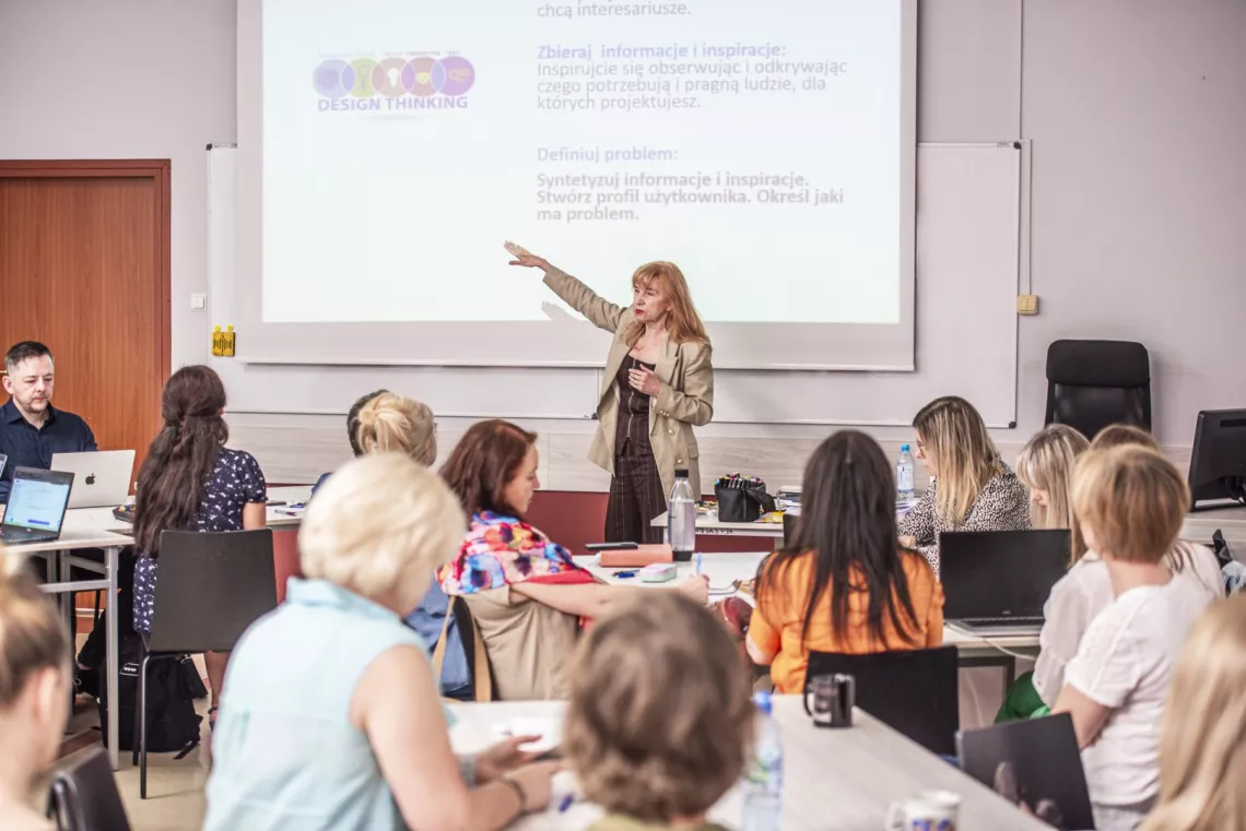 Kobieta prowadzi warsztat z metody Design Thinking. Stoi przy ekranie, wskazując na slajd z prezentacją. Uczestnicy, siedzący przy stołach z laptopami i notatkami.
