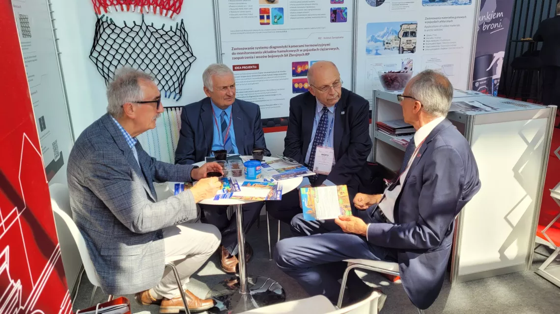 Czterech mężczyzn prowadzi dyskusję przy okrągłym stoliku, na ścianach stoiska wystawienniczego widoczne plakaty i inne elementy.