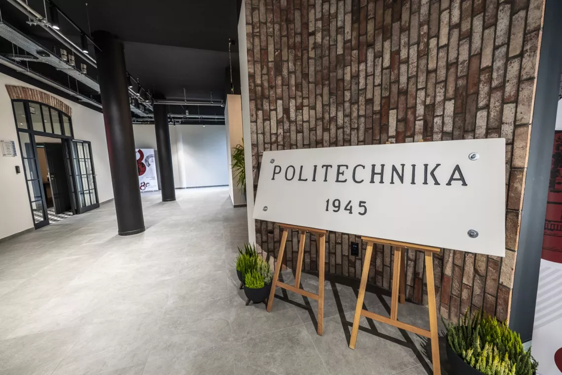 Tablica z napisem Politechnika 1945 na tle ceglanej ściany, widoczny korytarz, na którym stoja czarne kolumny