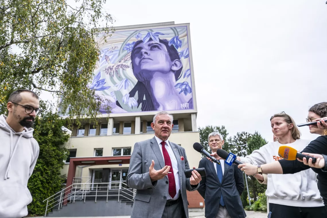 W czasie briefingu prasowego, stoi kilka osób jena z nich udziela wywiadu, w tle ściana z namalowanym na niej muralem