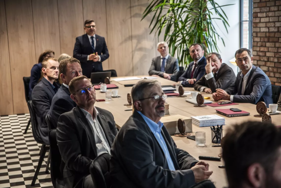 Mężczyzna stoi u szczytu stołu, przy którym sidzi kilkanaście osób i słucha jego prezentacji, patrząc na niewidoczny na zdjęciu ekran.