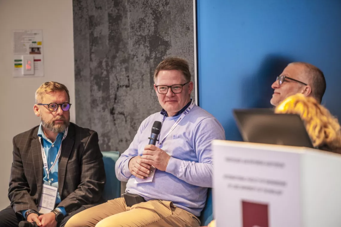 Trzej mężczyzni - uczestnicy panelowej dyskusji, jeden z nich trzyma mikrofon w ręku