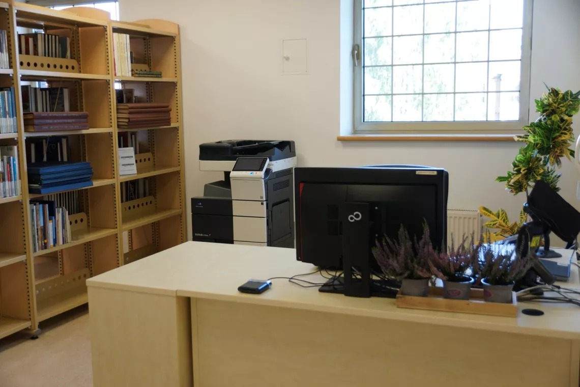 Stanowisko bibliotekarza dyżurującego, biurko z komputerem, obok regały i drukarka, w tle okno