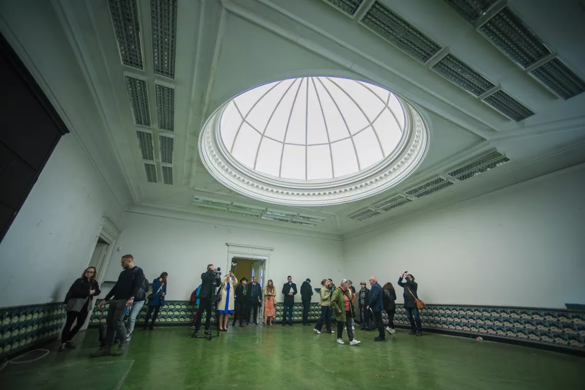 Duża sala bez mebli ze szklaną kopułą w suficie. Stoi w niej grupa kilkunastu osób.