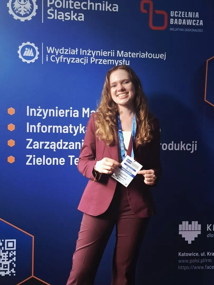 Uśmiechnięta młoda dziewczyna z długimi jasnymi włosami, trzyma identyfikator w ręku, stojąc na tle ścianki reklamującej wydarzenie