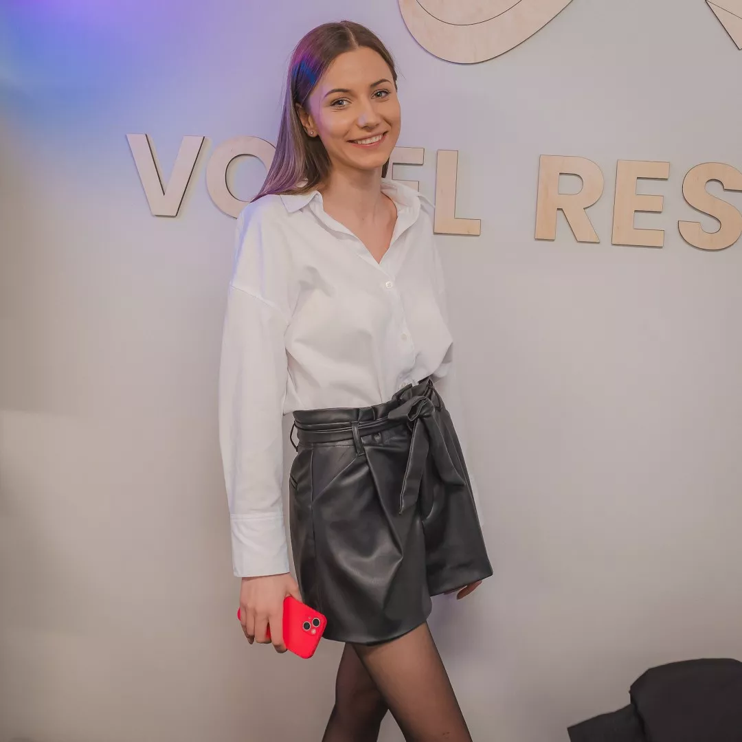 Młoda uśmiechnięta kobieta w bialej bluzce i czarnej spódniczce stoi na tle częsciowo widocznego napisu Voxel Research Lab