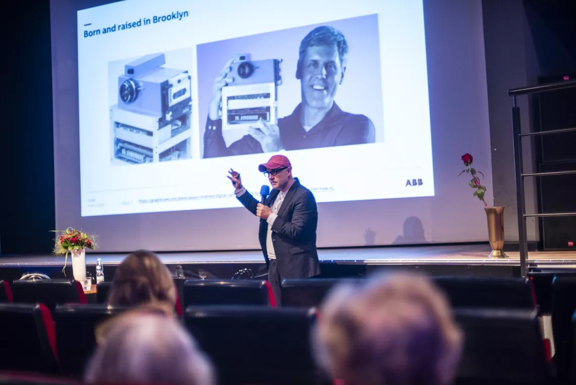 Mężczyzna z mikrofonem w ręku stoi przed publicznością, w tle ekran, na którym widać mężczyznę trzymającego jakieś urządzenie