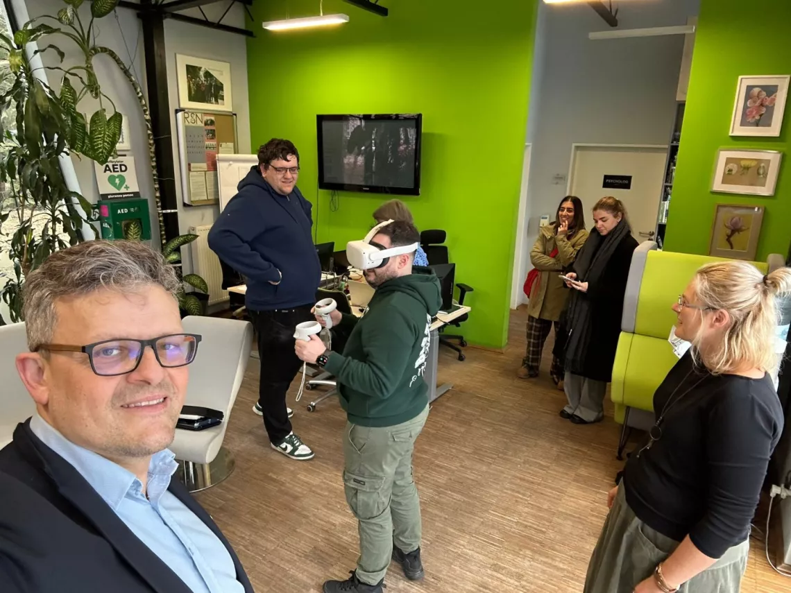 Kilkoro osób stoi w pokoju z zielonymi scianami, na których wiszą obrazki i telewizor,  