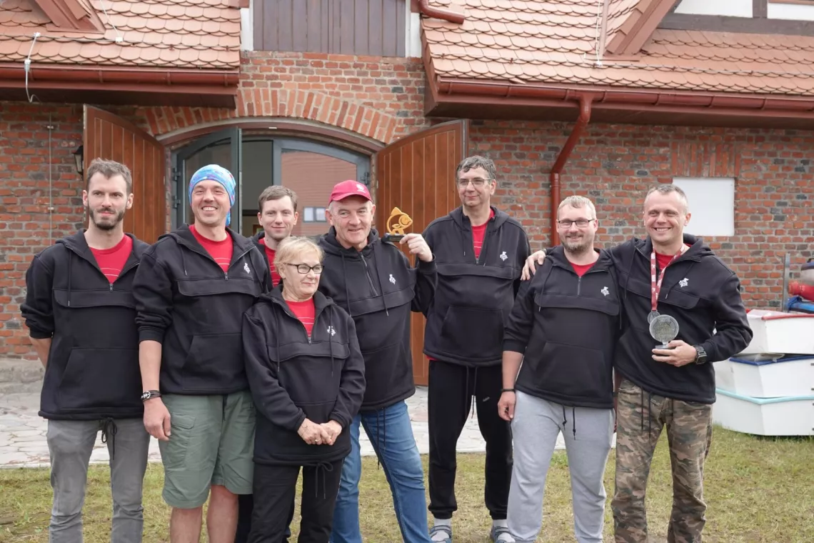 Grupa osób w czarnych bluzach stoi przed wejściem do budynku z cegły, pokrytego czerwoną dachówką