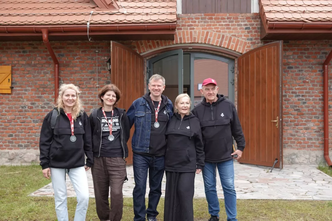 Trzy kobiety i dwóch mężczyzn z czarnych bluzach - troje ma medal na szyi, stoja przed wejściem do budynku z cegły, pokrytego czerwoną dachówką