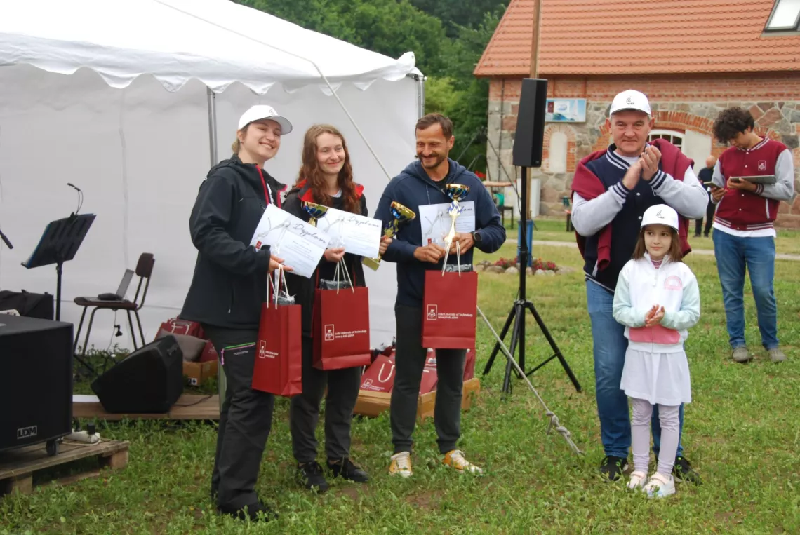 Trzy osoby stoją z pucharem i dyplomami w rękach, obok męższyzna i mała dziewczynka biją brawo, w tle namiot i budynek z czerwonej cegły
