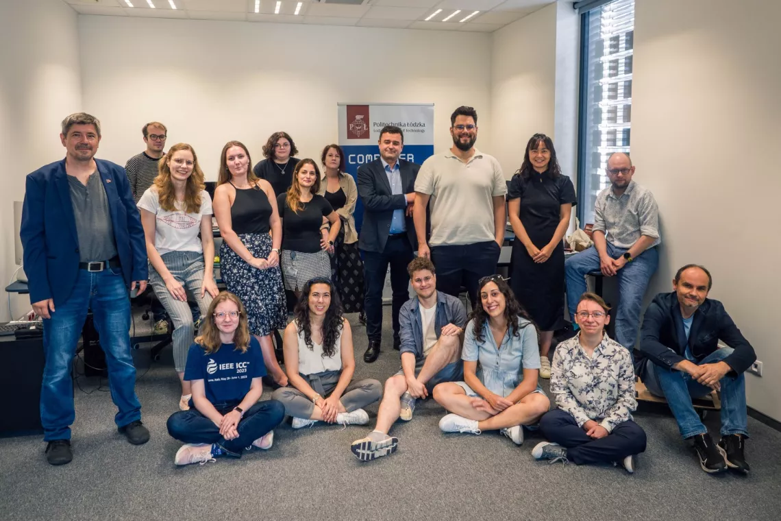 Grupa 17 osób pozuje do zdjęcia w sali, w tle rollup, po prawej okno. Część osób stoi, część siedzi na podłodze.