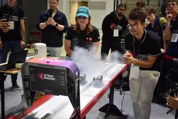 Młodzi ludzie przy torze, na którym widać dym od rozpedzonego autka, wokół publiczność obserwująca wydarzenie