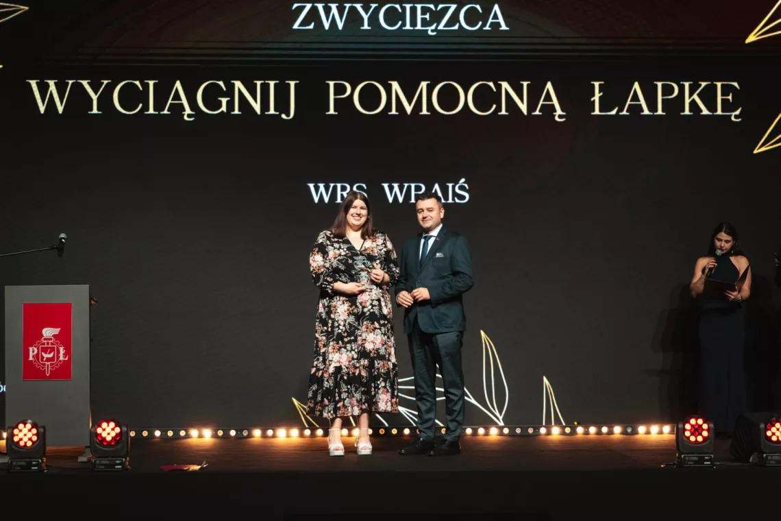 Kobieta trzymająca statuetkę i mężczyzna stoja na tle napisu zwycięzca Wyciągnij pomocną łapkę