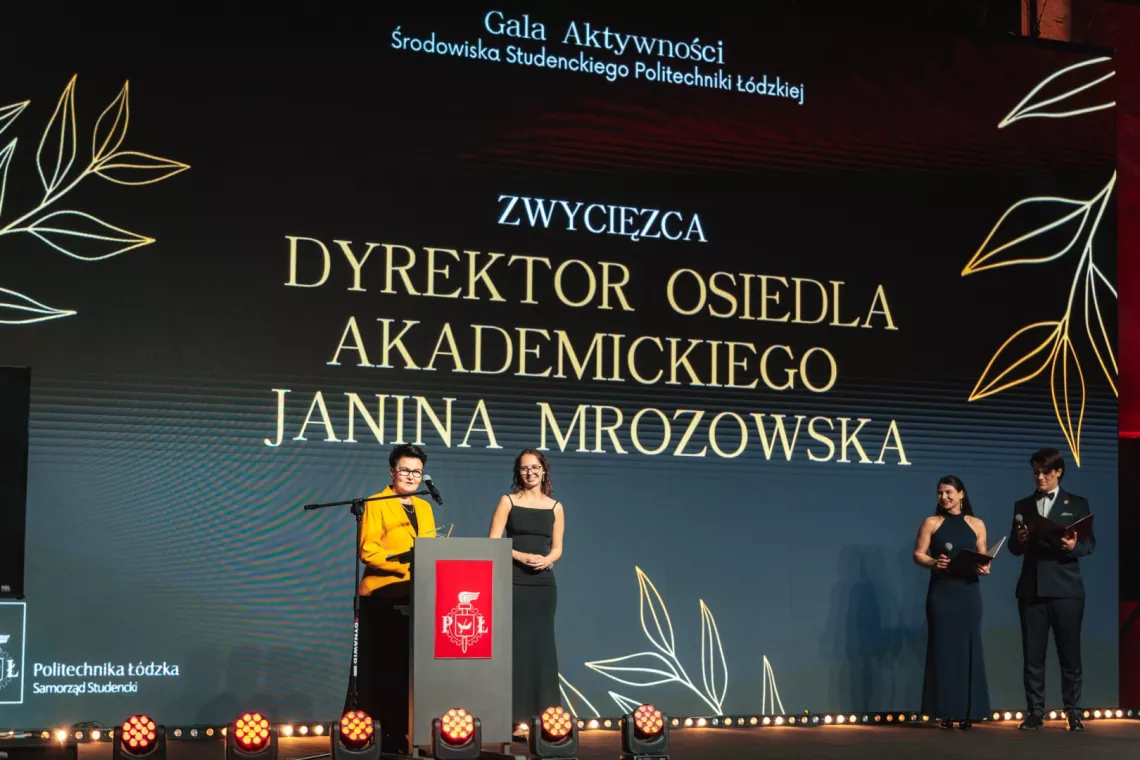 Kobieta w żóltej marynarce przemawia do mikrofonu, obok stoi dziewczyna w długiej, czarnej sukni. W tle ekarn z napisem Zwycięzca dyrektor osiedla akademickiego Janina Mrozowska