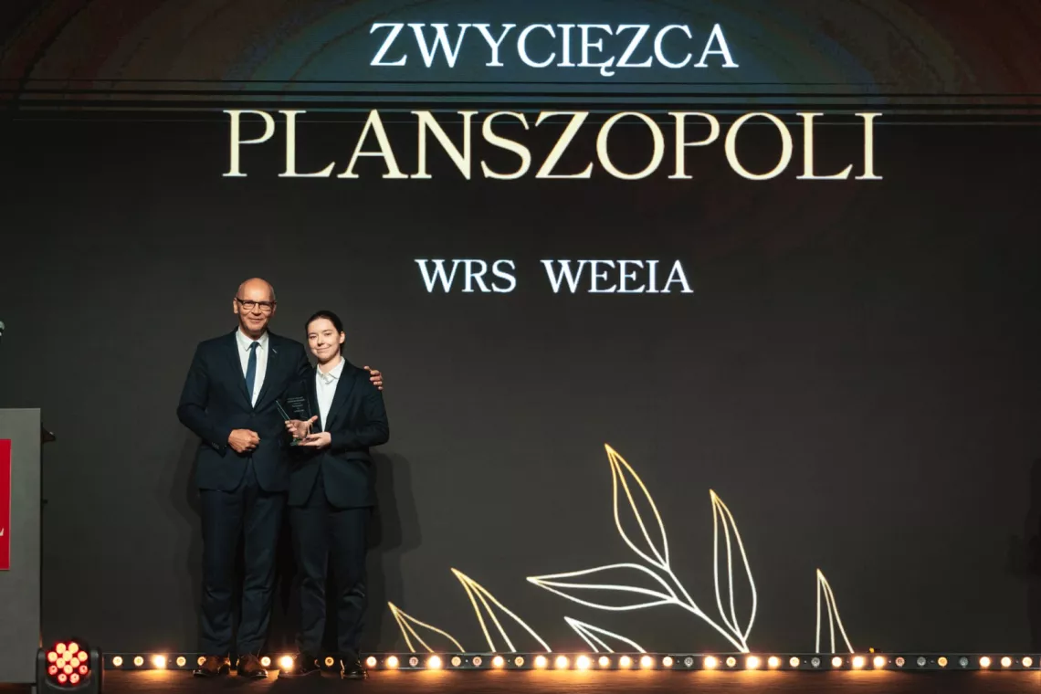 Kobieta trzymająca statuetkę i mężczyzna stoją na tle napisu zwycięzca Planszopoli