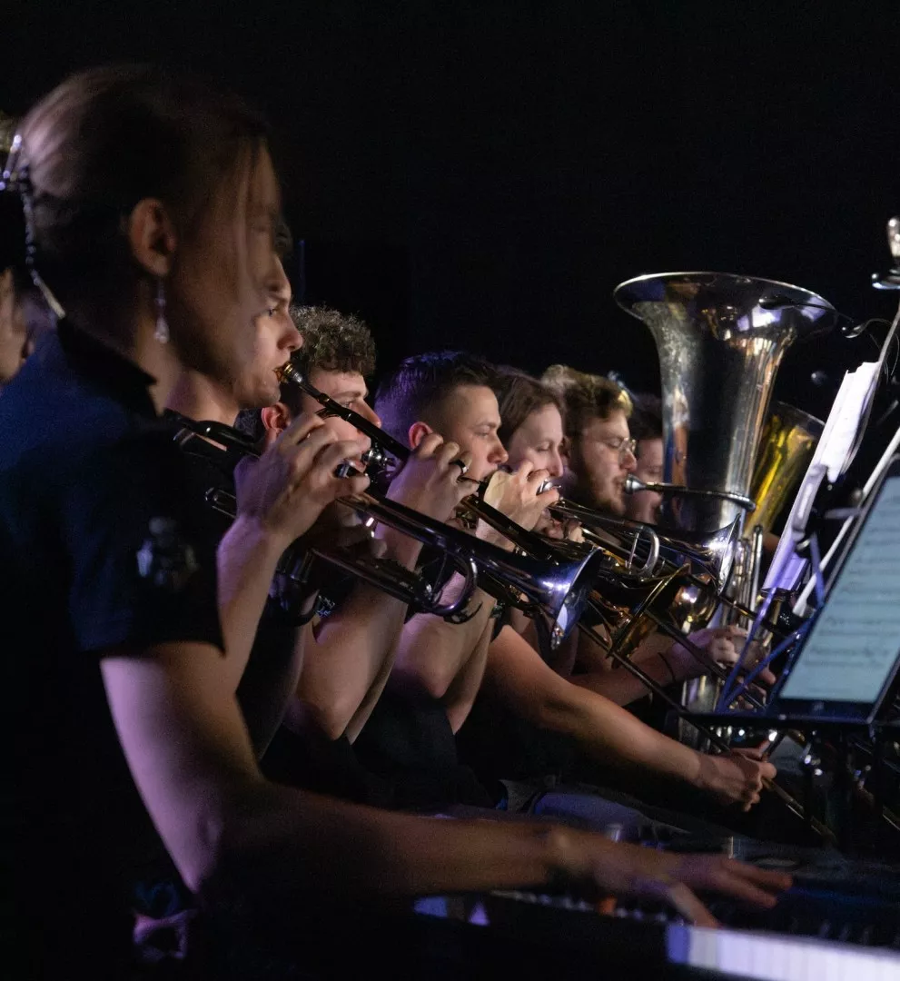 Grupa muzyków grających na instrumentach dętych blaszanych, takich jak trąbki i tuba oraz pianistka w ustawieniu orkiestralnym.Tło jest ciemne, co podkreśla wykonawców i ich instrumenty.