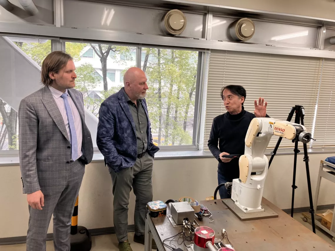 Trzy osoby stojące wokół stołu z różnym sprzętem, w tym z ramieniem robotycznym. Jedna z osób wyjaśnia lub demonstrować coś związanego z ramieniem robotycznym pozostałym dwóm. Sceneria wygląda na laboratorium lub warsztat z dużymi oknami w tle.