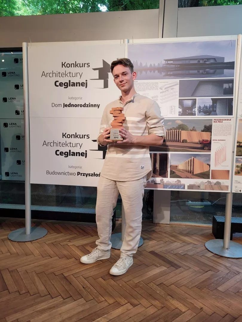 Młody mężczyzna trzyma w reu nagrode w formie nierówno ułożonych cegieł jedna na drugiej. Z tyłu planasza z napisem Konkurs Architektury Ceglanej.