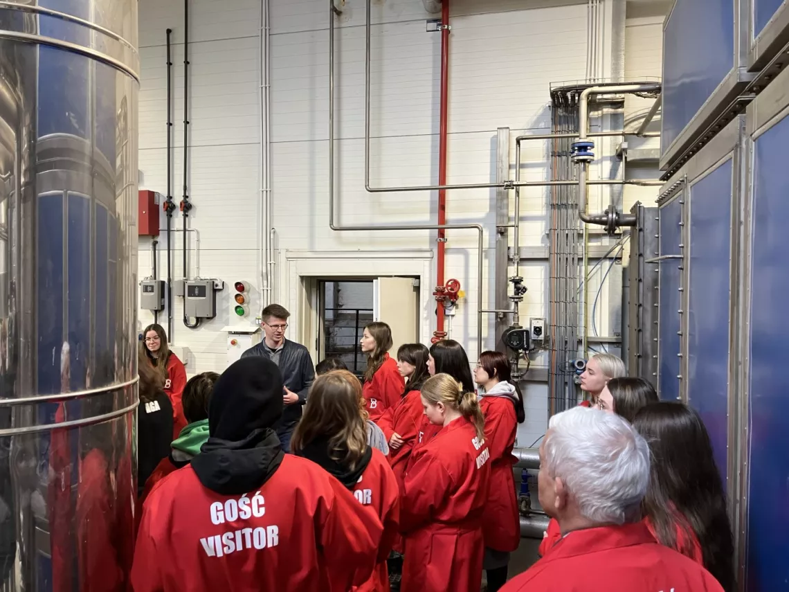 Grupa osób w czerwonych fartuchach z napisem visitor słucha prelekcji  w sali o charakterze przemysłowym