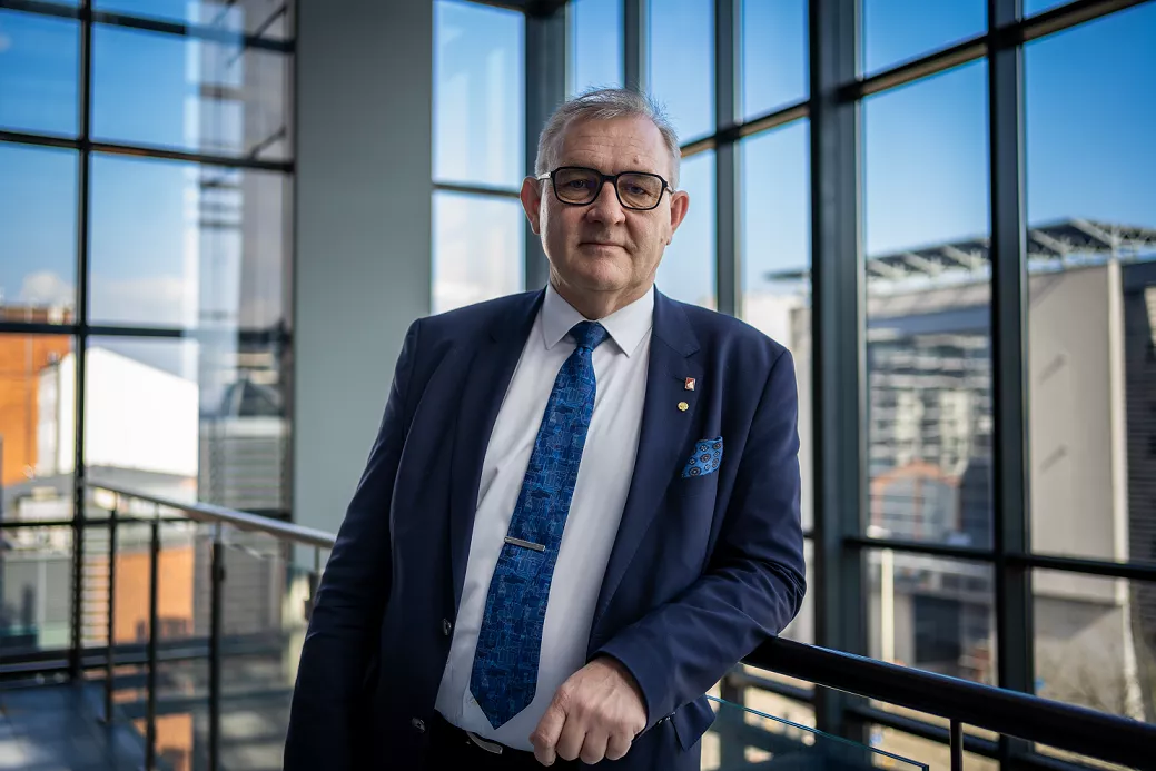 Mężczyzna w formalnym ubraniu stoi na tle opzreszkolen ściany, oparty o poręcż (ujęcie postaci w planie średnim)