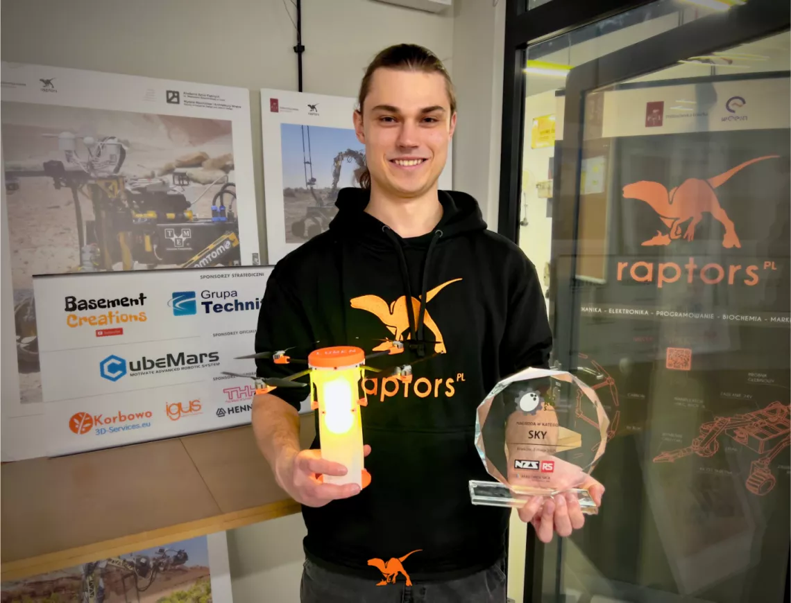 Osoba w bluzie z napisem raptors trzyma w ręku dron i nagrodę w formie szklanej plakiety