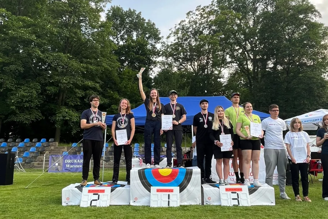 Zawodnicy na podium, w ręku trzymają dyplomy i mają medale zawieszone na szyi. W tle namiot i drzewa.