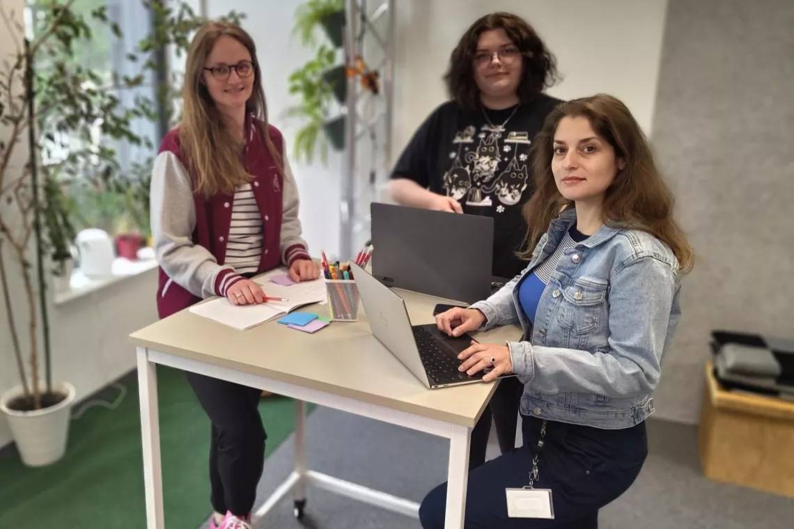 Trzy kobiety stoją przy stole, na którym leżą laptopy i kartki. Z lewej strony jest okno i doniczki z roślinami.