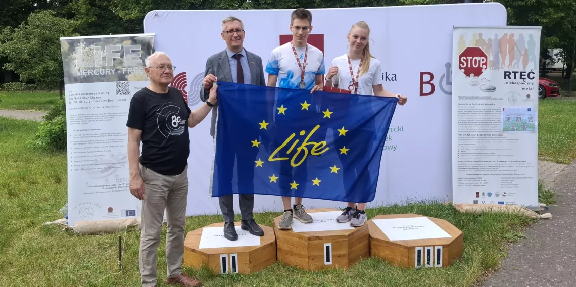 Dwoje młodych ludzi stoi na podium razem z elegancko ubranym mężczyzną, trzymają flagę unijną z napisem life, obok stoi jeszcze jeden mężczyzna
