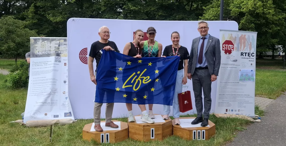 Trzy kobiety w sportowych strojach i dwaj mężczyżni stoją na podium, wspólnie trzymają flagę unijną z napisem life