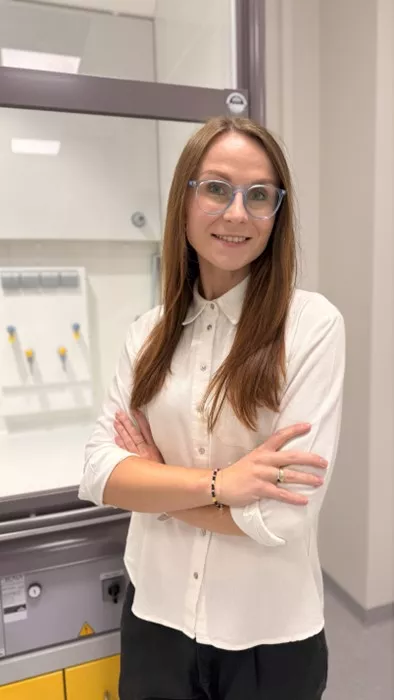 Dr inż. Magdalena Gajek stoi z założonymi rękami w laboratorium. Ma na sobie białą koszulę i okulary, uśmiecha się do obiektywu. W tle widoczne są elementy wyposażenia technicznego.