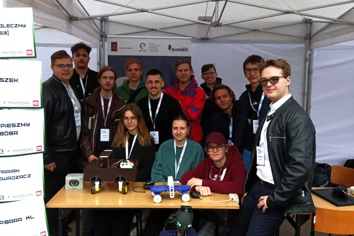 Kilkanaście osób pod namiotem wystawiennicznym, skupieni wokół stołu na którym leżą rózne urządzenia